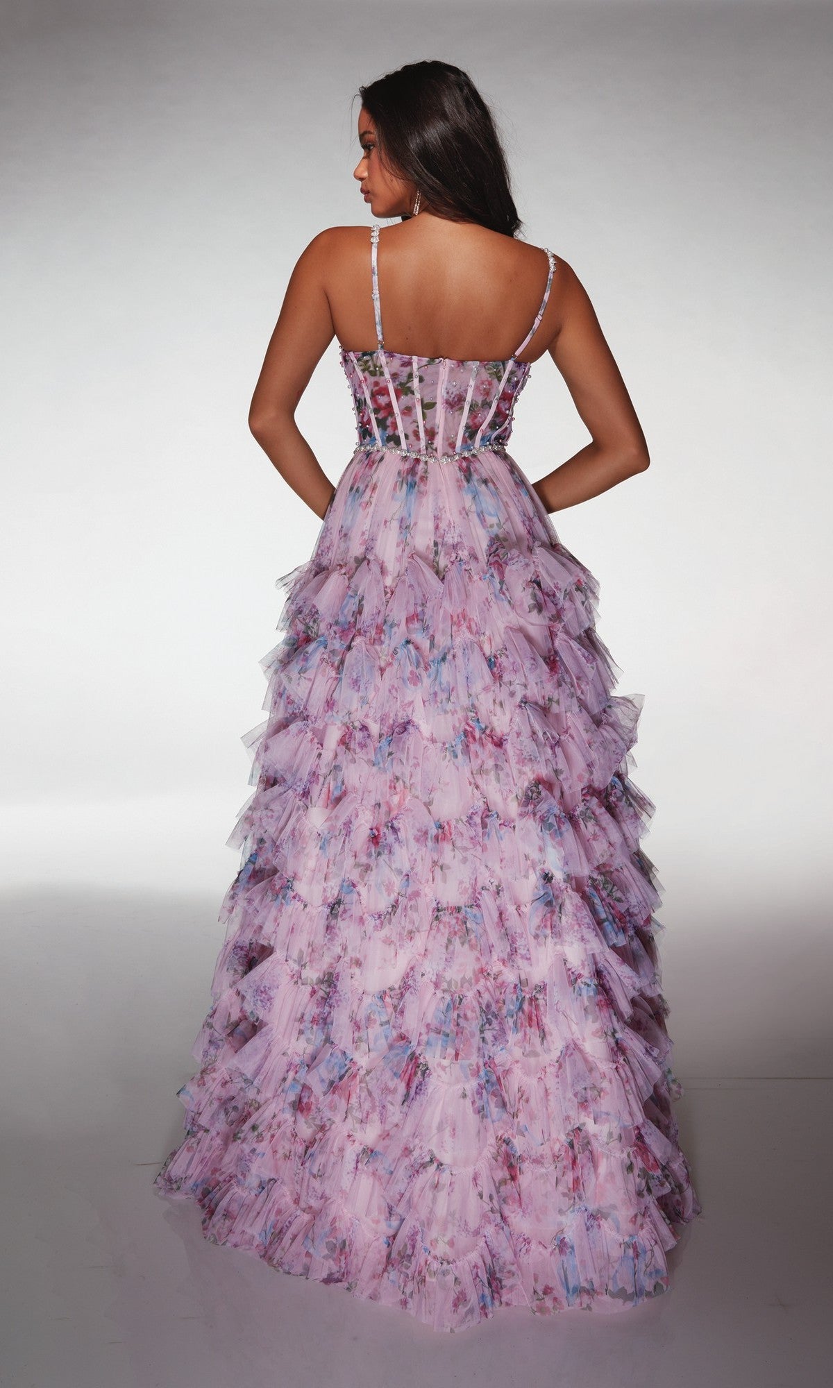 A woman facing away wears the Alyce 62182 long prom dress, featuring a sleeveless sheer corset bodice and a tiered ruffled skirt in a pink floral pattern, set against a plain background.